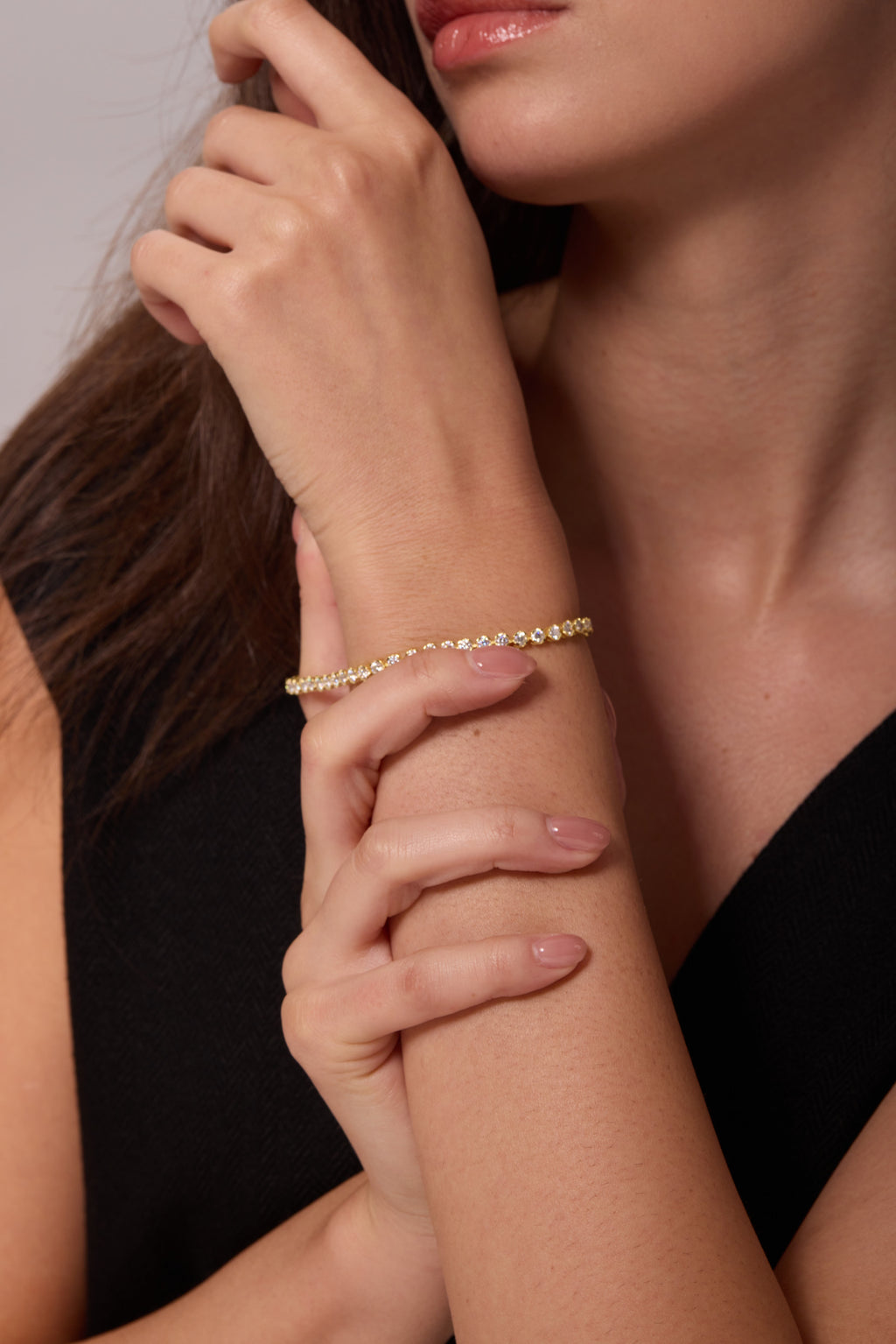 Gold-tone bracelet with a row of round clear stones, worn on a raised wrist supported by the other hand.