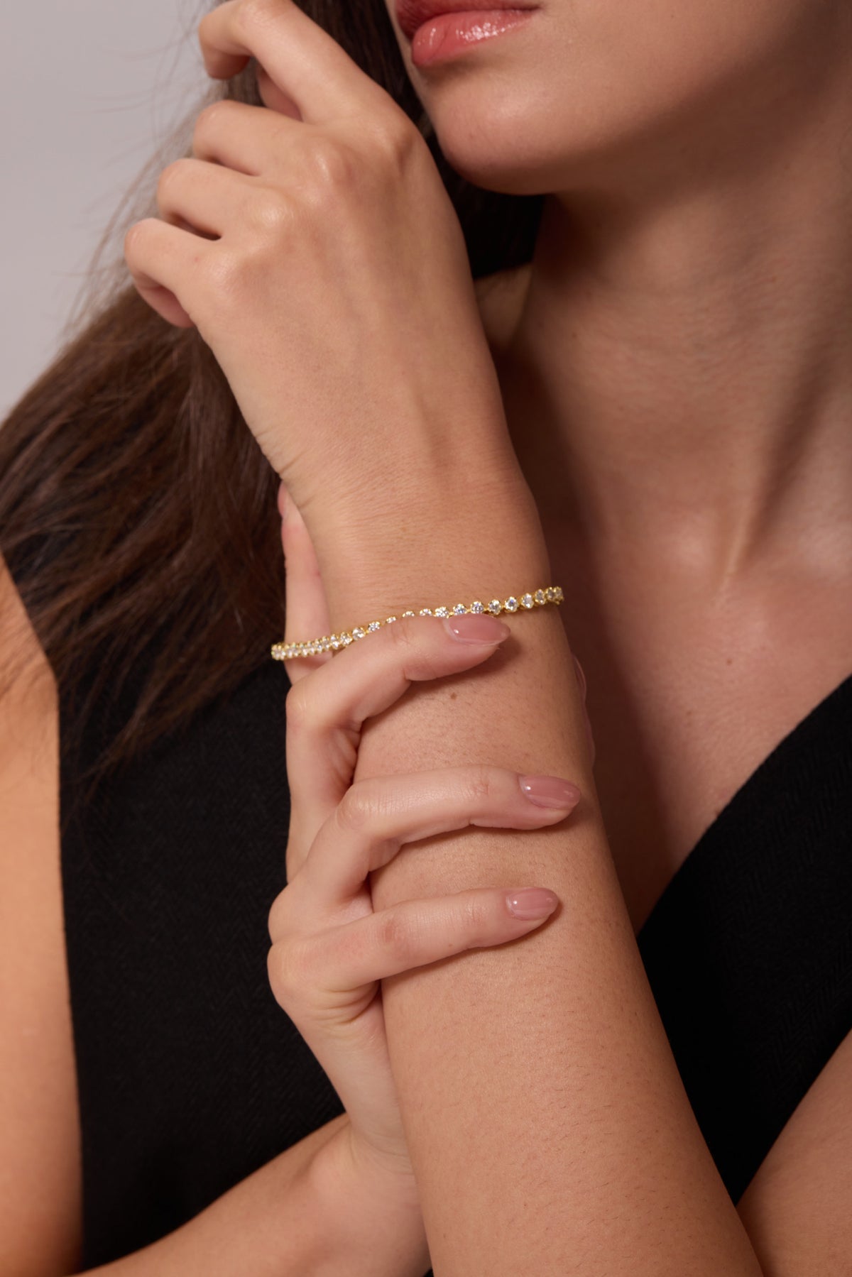 Gold-tone bracelet with a row of round clear stones, worn on a raised wrist supported by the other hand.