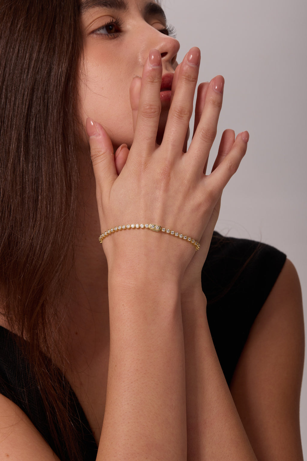 Gold-tone bracelet with round clear stones on a raised wrist, with the hands held near the lips.