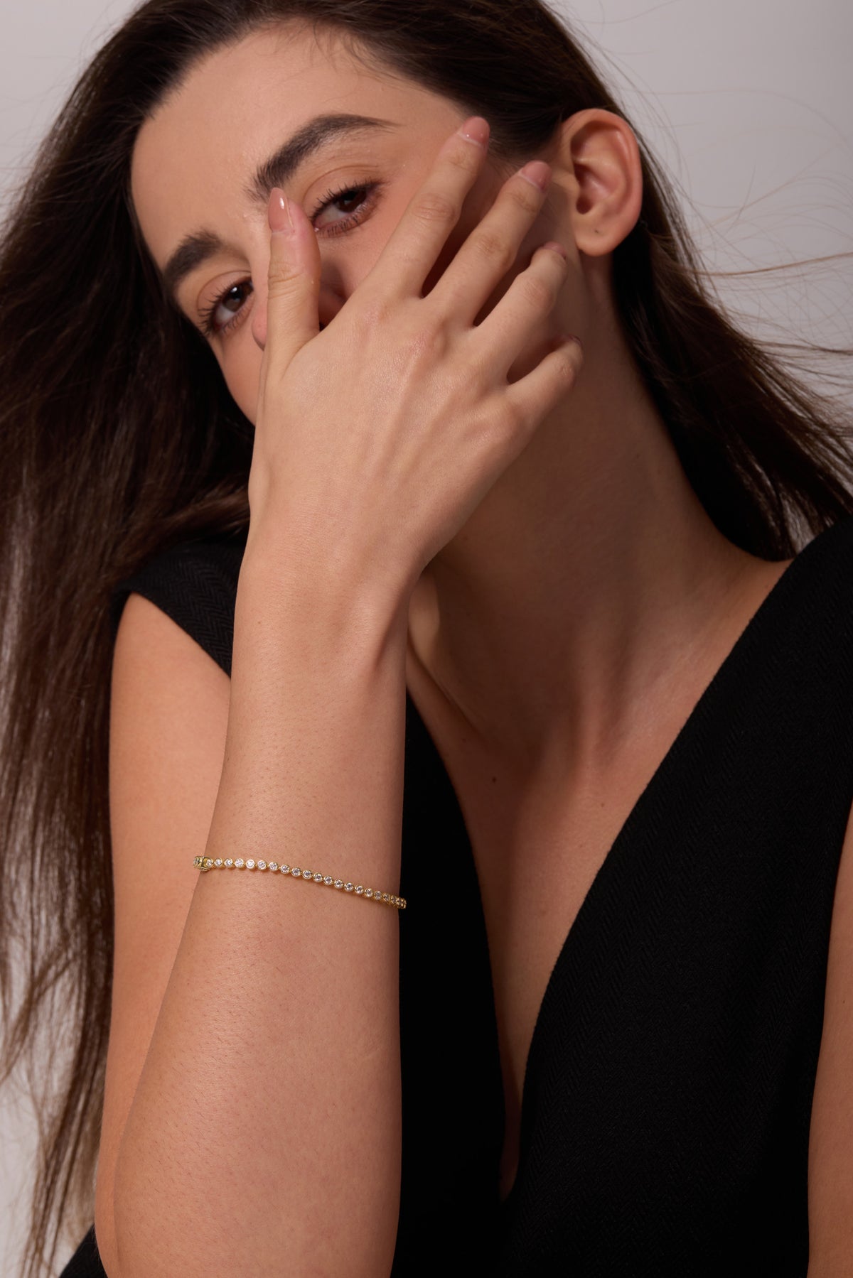 Gold-tone bracelet with round clear stones on a raised wrist beneath a hand resting across the cheek.