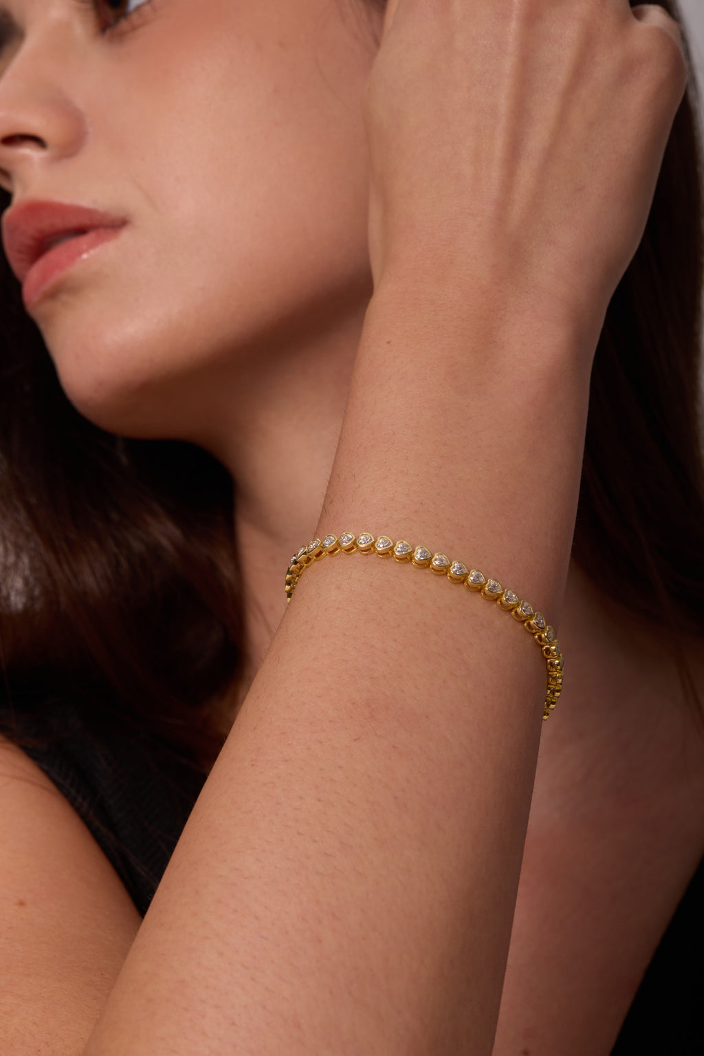 Close-up of heart-shaped clear stones in gold-tone bracelet settings on a raised wrist beside the face.