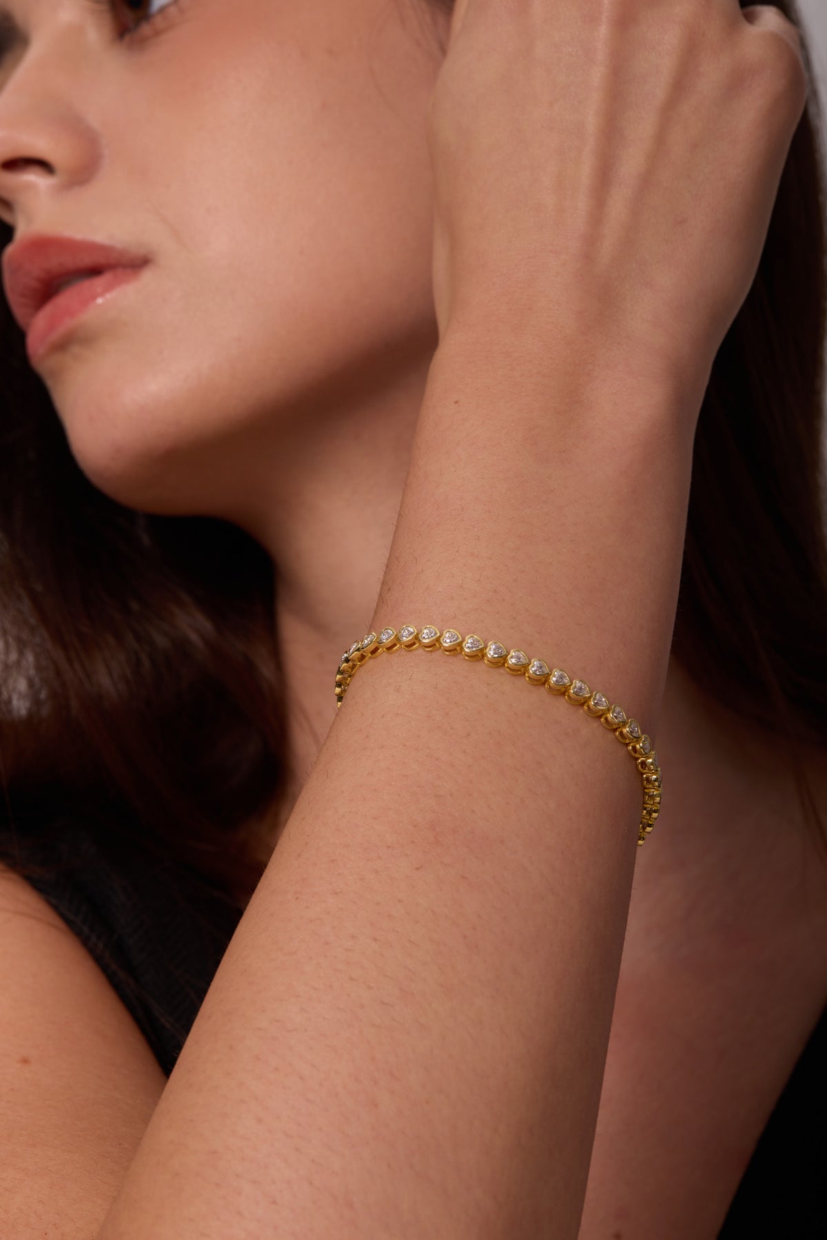 Close-up of heart-shaped clear stones in gold-tone bracelet settings on a raised wrist beside the face.