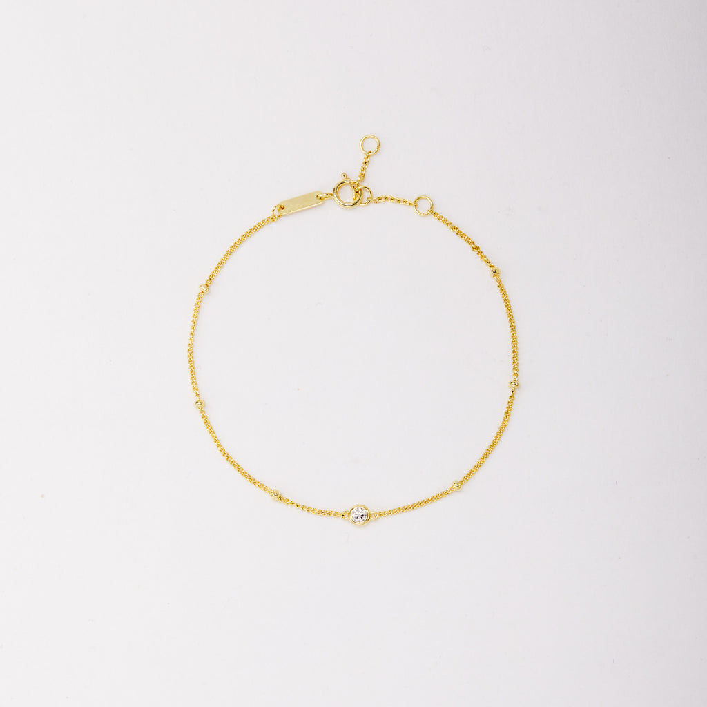 Gold-tone chain bracelet with small beads and a single round bezel-set stone, shown laid flat on a light background.