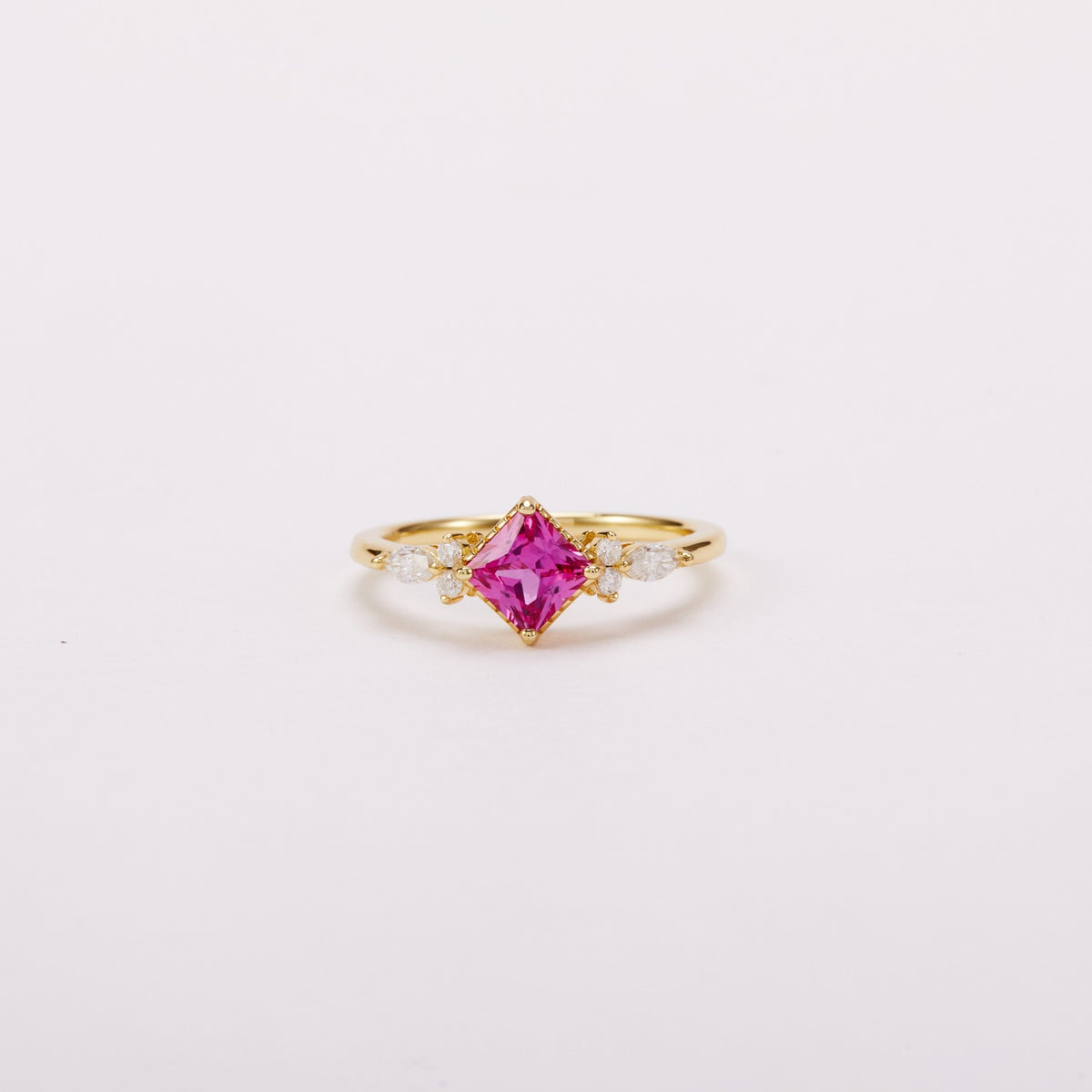 Front view of a gold-tone ring with a vivid pink square center stone set diagonally and clear wing-shaped side clusters.