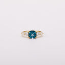 Front view of a gold-tone ring with a teal cushion-shaped center stone and four clear side stones.