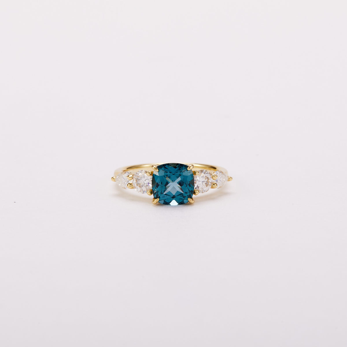 Front view of a gold-tone ring with a teal cushion-shaped center stone and four clear side stones.